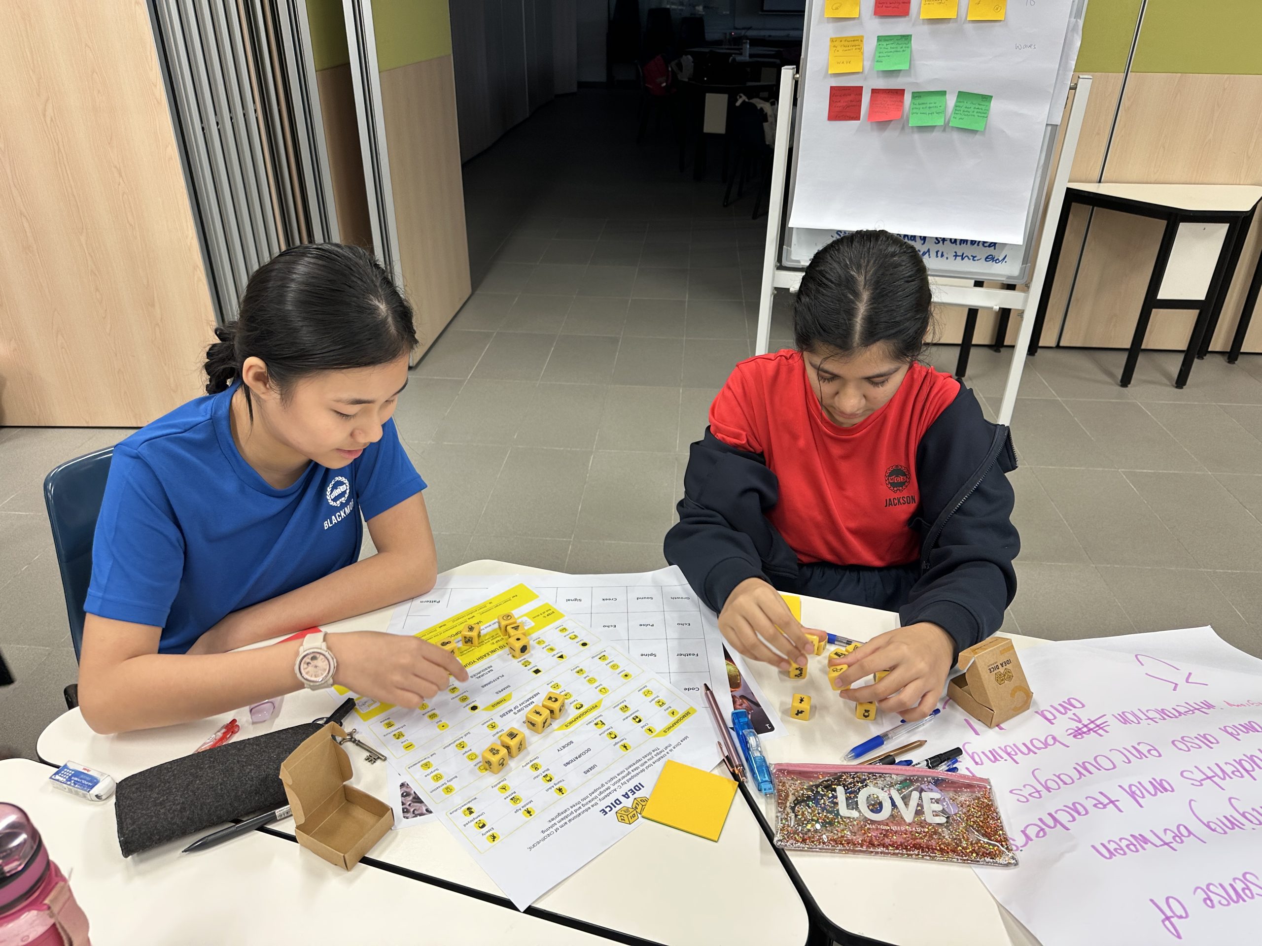 Two children sit at a table working with yellow dice and worksheets in a classroom setting.